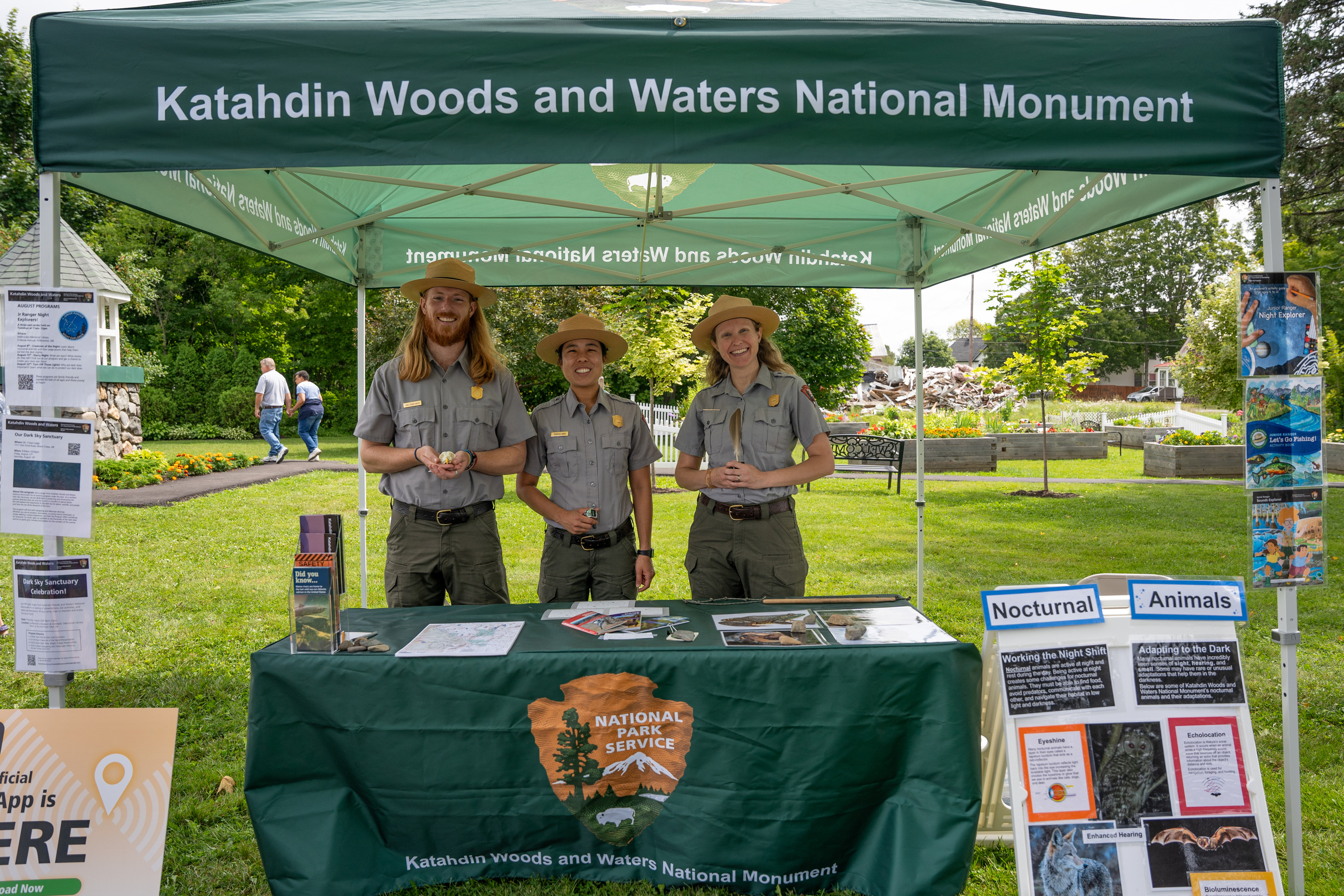 A green shade tent is set up over a table with the National Park Service arrowhead. Both the tent and table read Katahdin Woods and Waters National Monument. There are posters and fliers set up on either side of the table and three rangers stand behind the booth in flat hats. 
