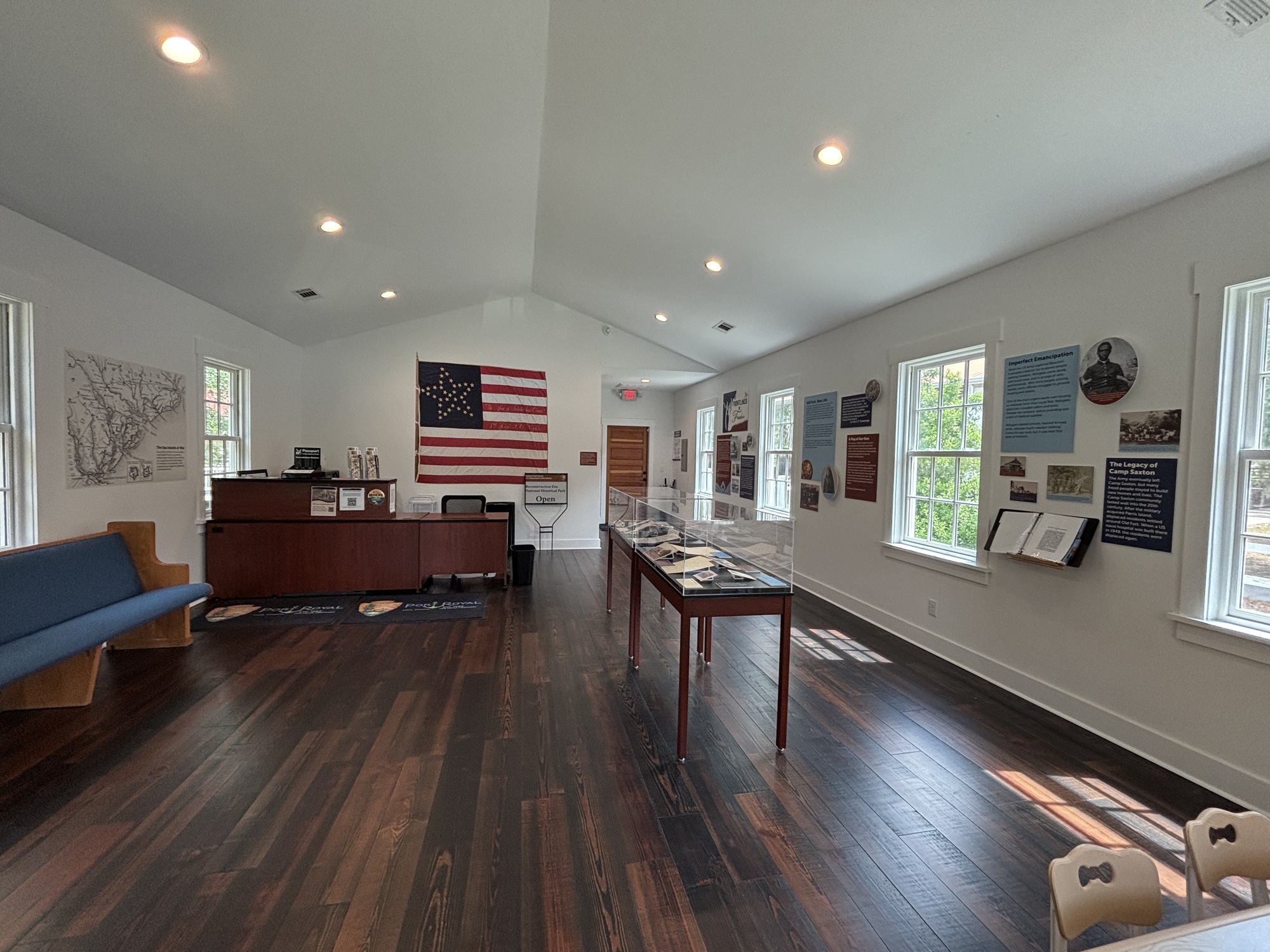 A large room with dark wooden floors and white walls. The left side of the room has a pew and information desk at the back with a large American flag on the wall. The right side has an exhibit along the wall and two exhibit cases closer to the middle of the room. Next to the door on the right is a small children’s table with coloring sheets.