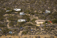 A view of a few campsites in a campground. Two roads cross through desert vegetation and there are about five vehicles and three tents. There is also a small tan and brown building. 
