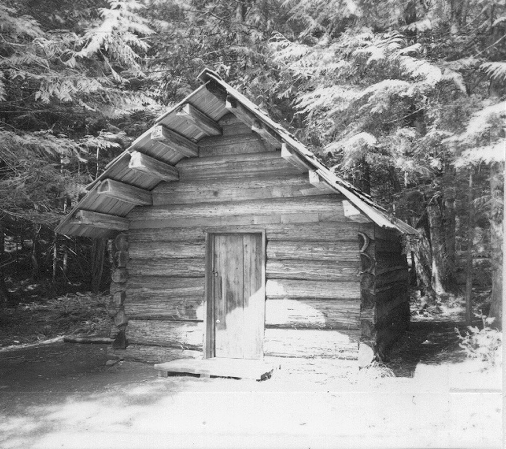 A small wood cabin with cedar plank sides and roof and a single door surrounded by forest. 