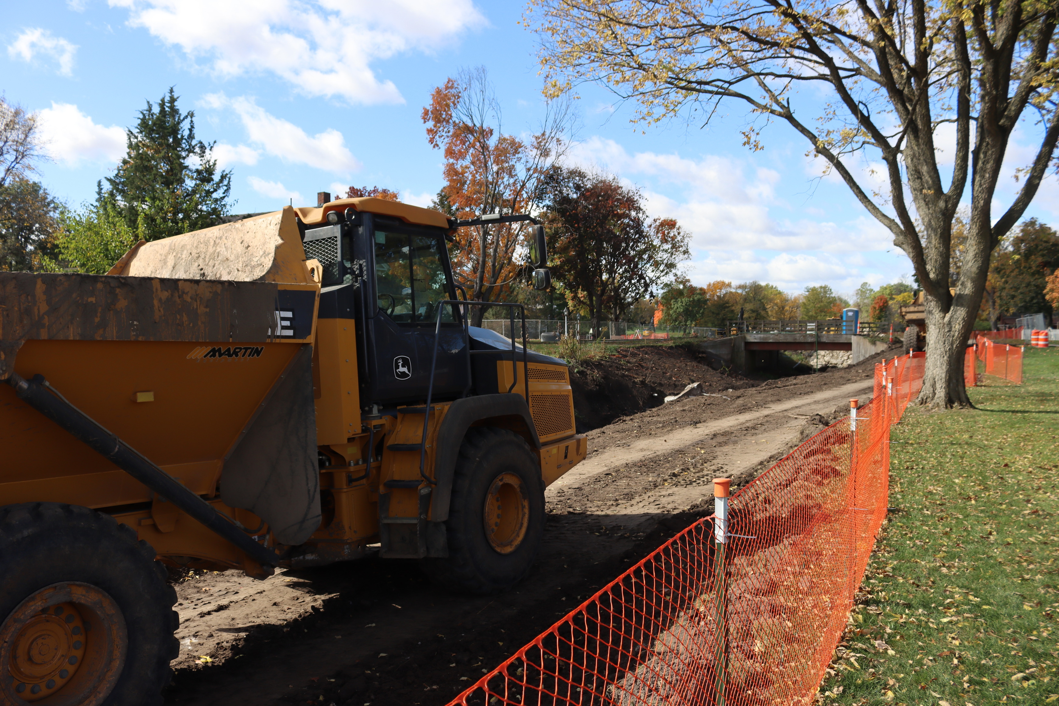 A large yellow dump truck is parked alongside the creek waiting to move into position for its next load. An orange fence runs alongside the rode from the bottom center of the fence into the distance.