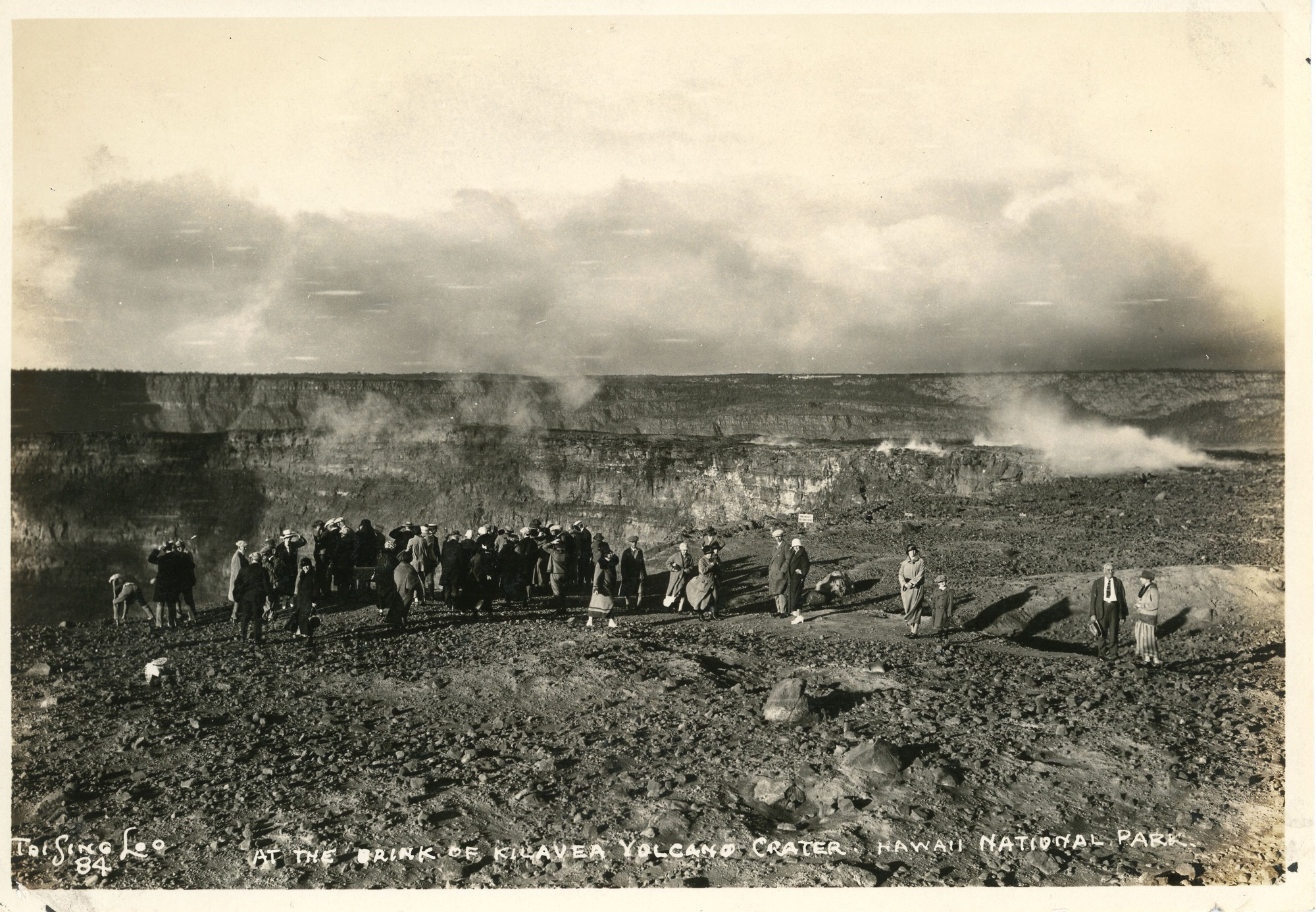 A black and white image of a large group of visitors standing at the brink of the Kīlauea volcano crater. The group of people standing at the edge of the crater appear to be men, women, and children. Most of them are standing at the edge of the crater with their backs towards the camera. Some people appear to be walking down the trail to the crater and others appear to be walking away from the crater. On the right side of the image there is a sign near the crater in the distance. The ground around the crater is rocky. In the background a wall of the crater is visible, and clouds are in the sky. On top of the wall there is a tiny glimpse of Volcano House.  On the bottom left corner of the image, there is a photographer signature, " Tai Sing Loo 84." The bottom of the image also has a caption that reads "At the brink of Kilauea Volcano Crater, Hawaii National Park."