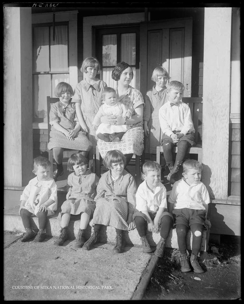 Mrs. McNulty and her ten children sitting on the front porch of a shingled house. 
