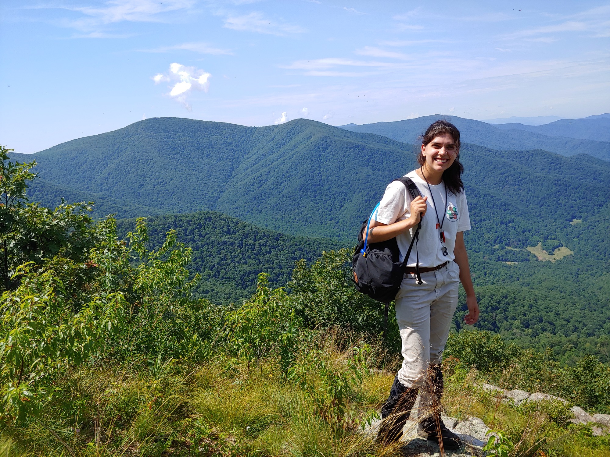 A woman in a tan t-shirt stands with a backpack over one shoulder at an overlook with green, tree-covered mountains in the background.