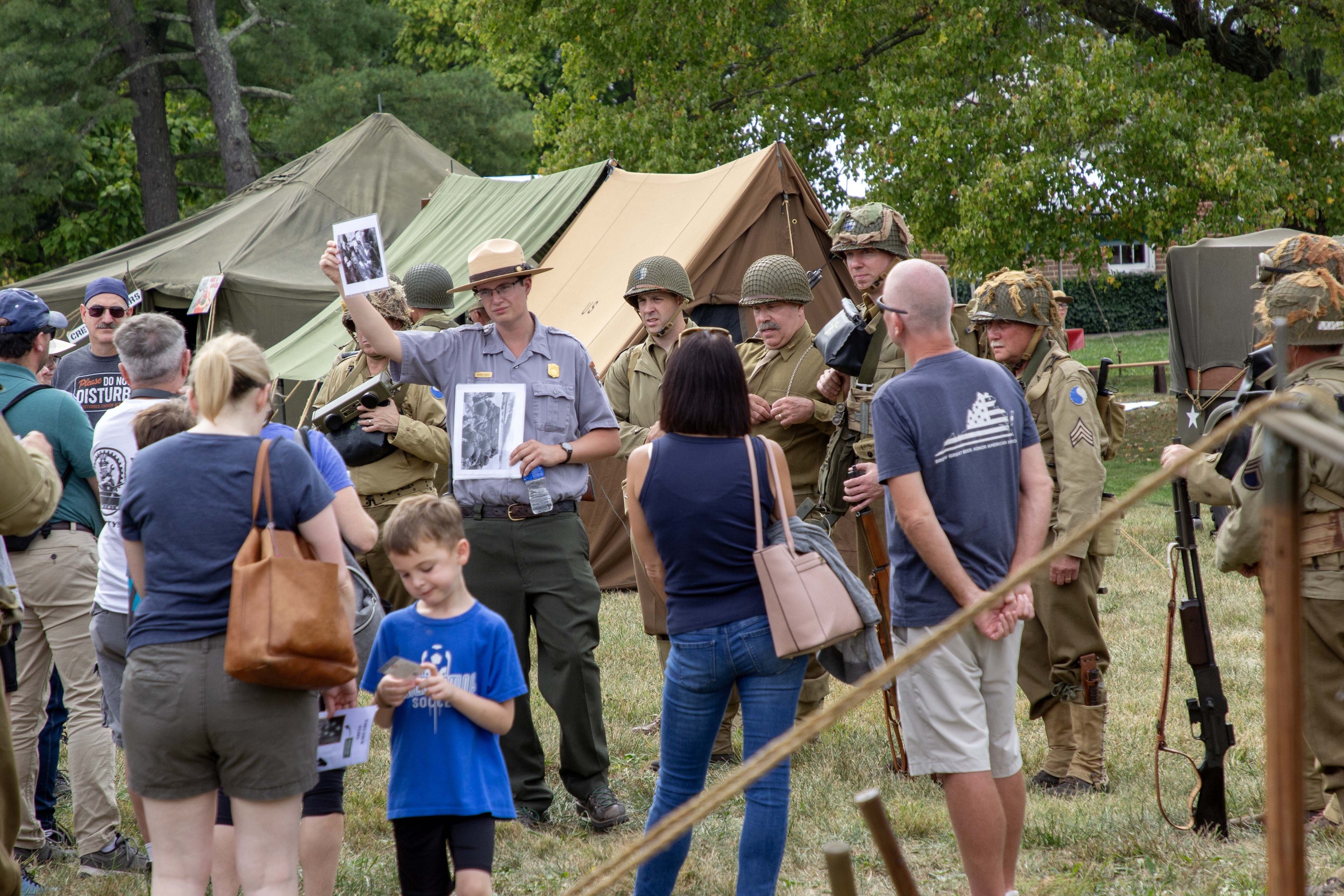 A group of visitors listen to a park ranger who is holding up pictures. There are tents around them and people wearing WWII era uniforms. 