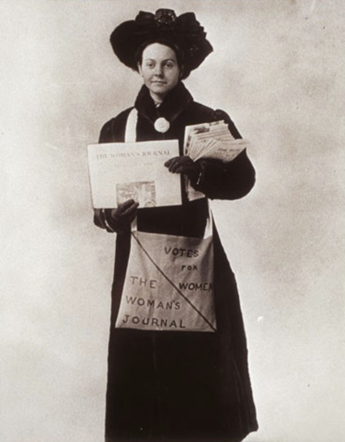 Florence Luscomb in a coat and hat standing, holding a copy of The Woman's Journal.