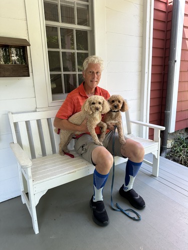 A man sits on a white bench on a porch holding two dogs.