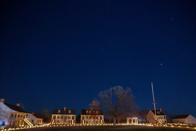 6 buildings, a small pavilion, and a flagpole are lit by candlelight from their paths in front and their front steps. Stars shine brilliantly in the sky.