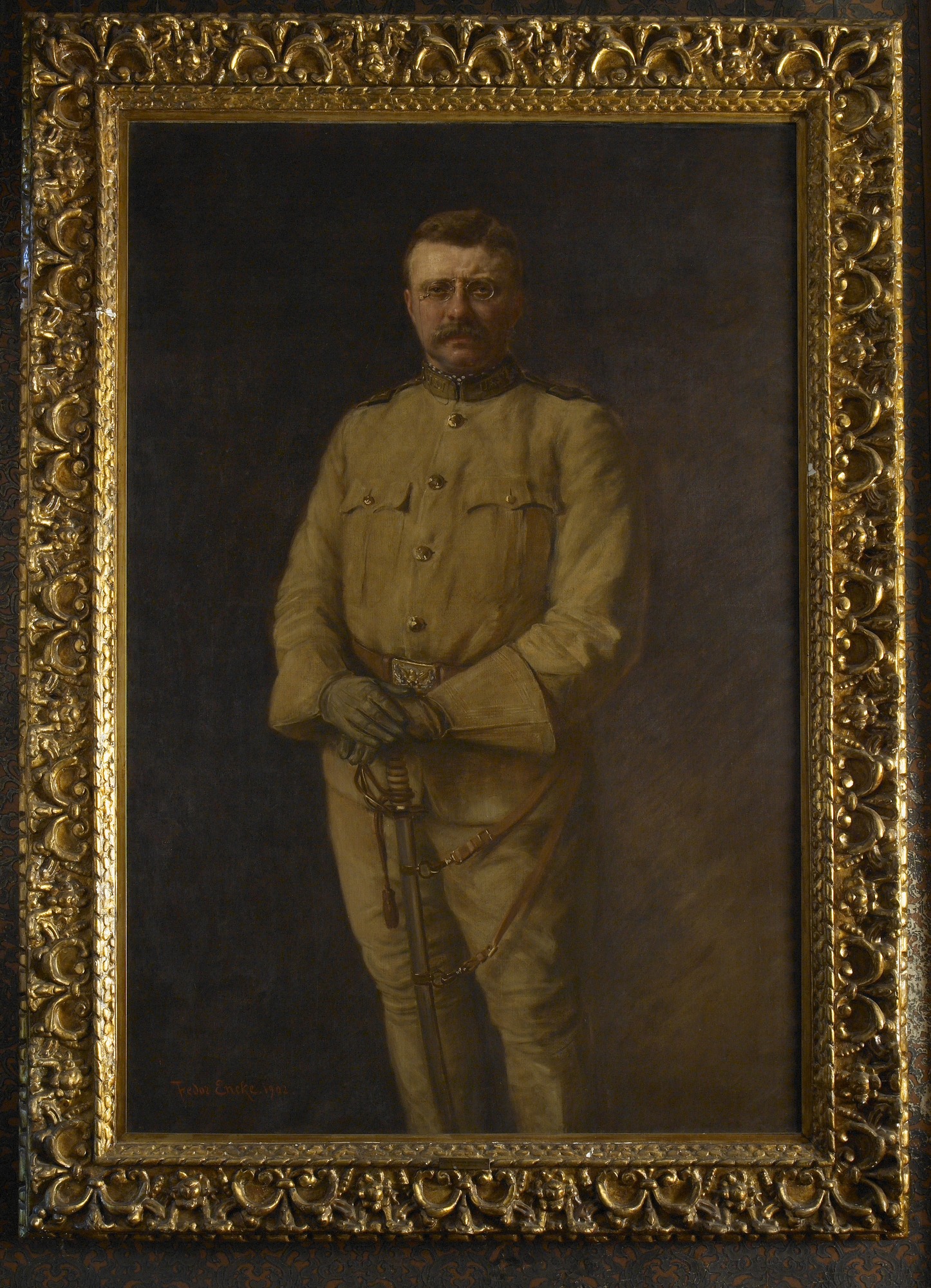 A portrait of Theodore Roosevelt standing and facing the viewer in his Rough Rider uniform. The uniform is golden khaki, with long sleeves, long pants, golden buttons, and a golden belt buckle. He wears gloves and holds his saber on the handles with both hands and the saber facing downwards. He has pince-nez glasses, a mustache, and a serious expression. 