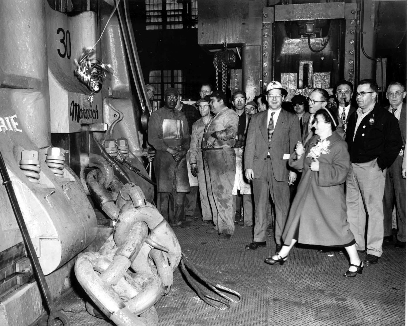 A large hammer in an industrial building surrounded my Navy Yard workers and some men in suits. A woman with flowers stands facing the hammer. 