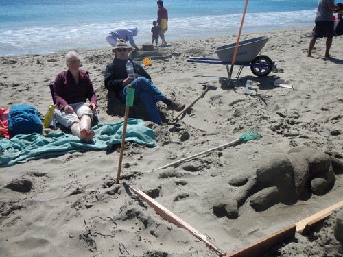 A woman and a man sitting beyond a sand sculpture of a dog with a bone.