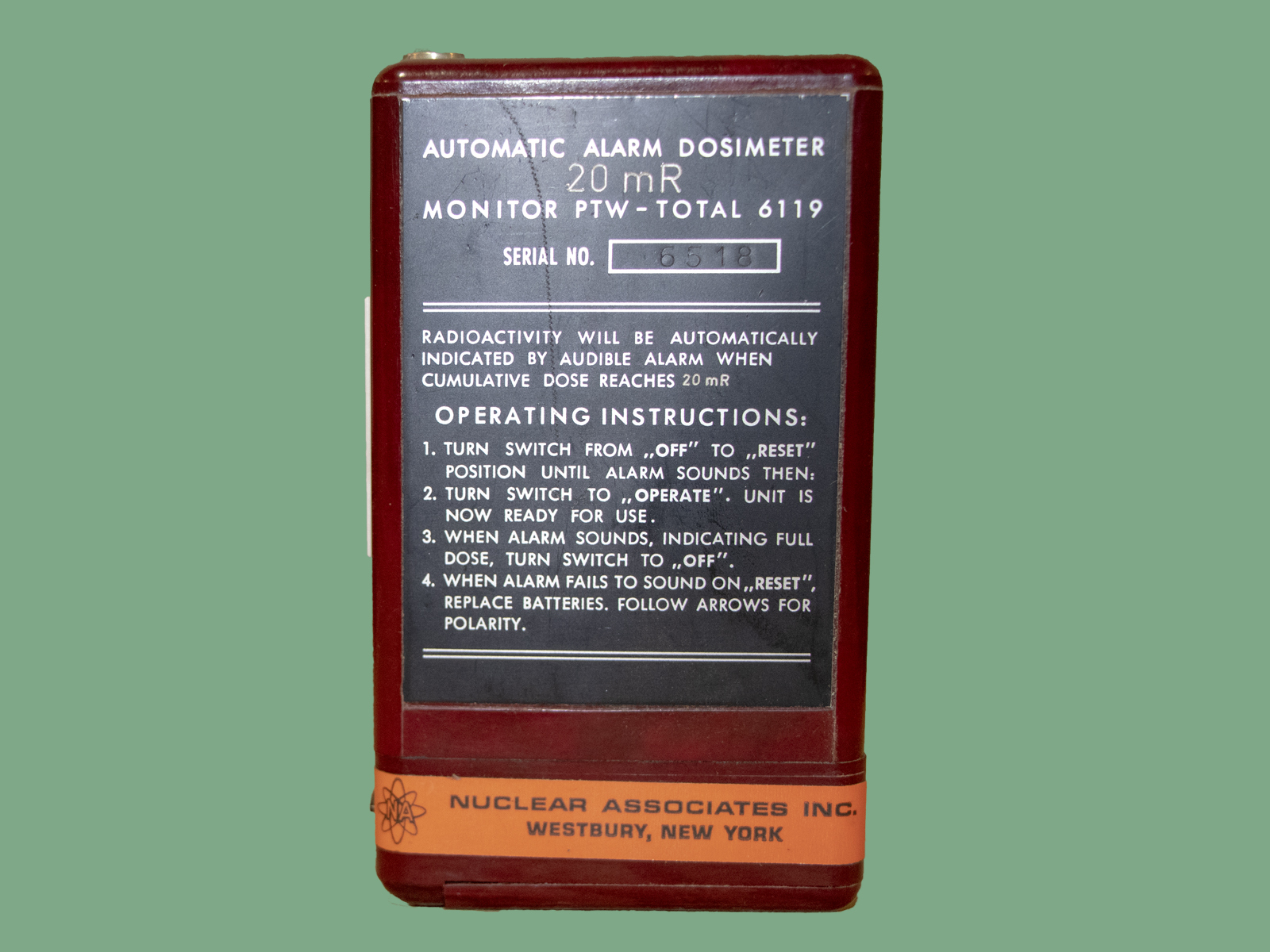 A red box with a black label that reads, "AUTOMATIC ALARM DOSIMETER 20mR MONITOR PTW-TOTAL 6119", "RADIOACTIVITY WILL BE AUTOMATICALLY INDICATED BY AUDIBLE ALARM WHEN CUMULATIVE DOSE REACHES 20 mR". This is followed by operating instructions. There is an orange label on the bottom that reads "NUCLEAR ASSOCIATES INC. WESTBURY, NEW YORK".