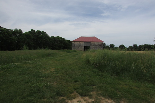 A rectangular stone barn has a tall opening in the center of one side and a hipped roof. A mowed path leads through tall grasses to the barn. 