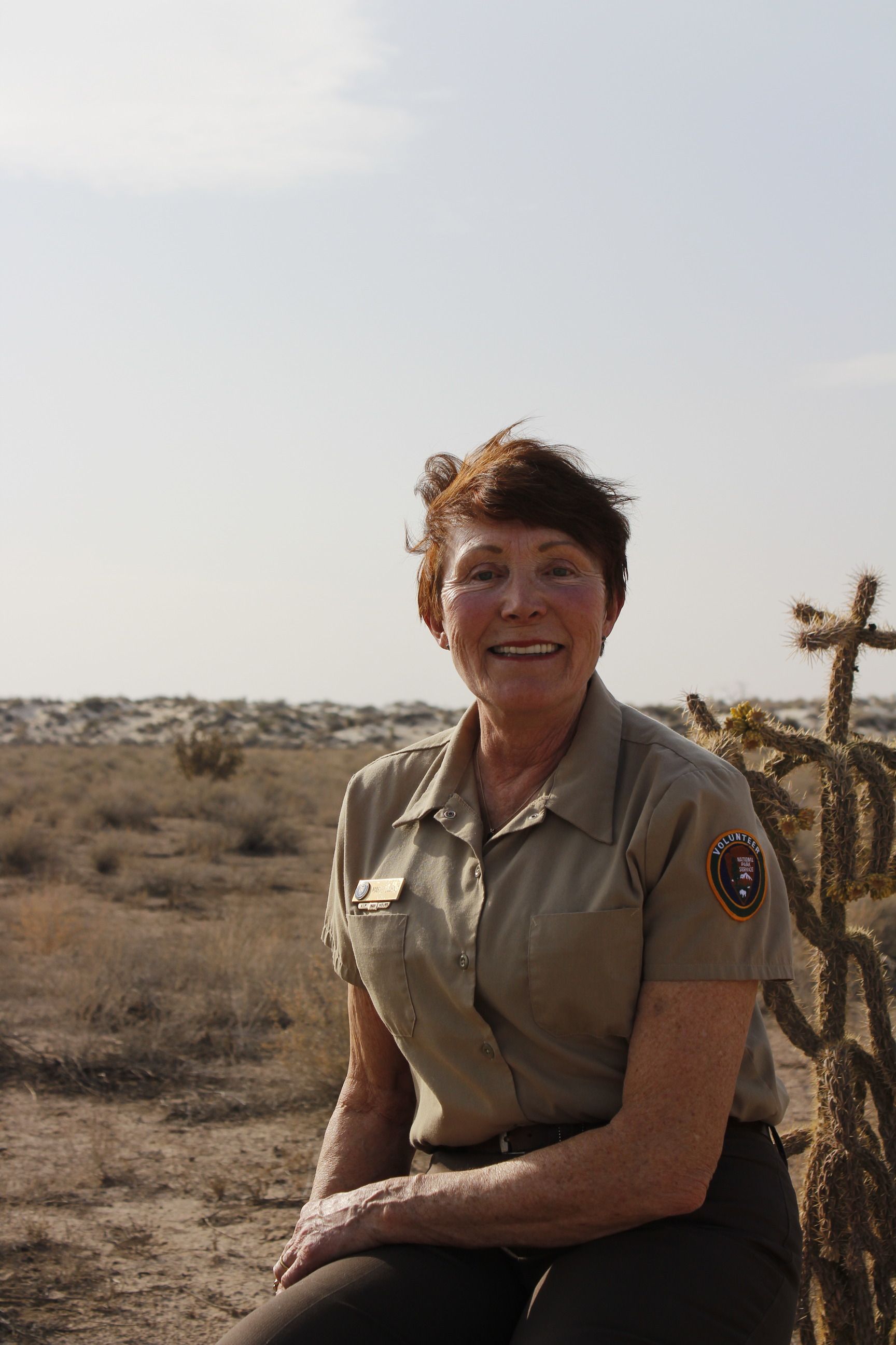 A woman proudly wearing the NPS volunteer uniform sits for a photo, smiling towards the camera. Behind her Cholla cactus and desert shrubbery unfold in the distance meeting white sand dunes freckled with plants. 