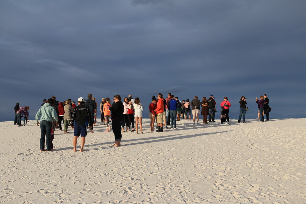 A large group of visitors attends the sunset stroll. The sun is shining from the west, but dark gray clouds look ominous in the eastern sky. 