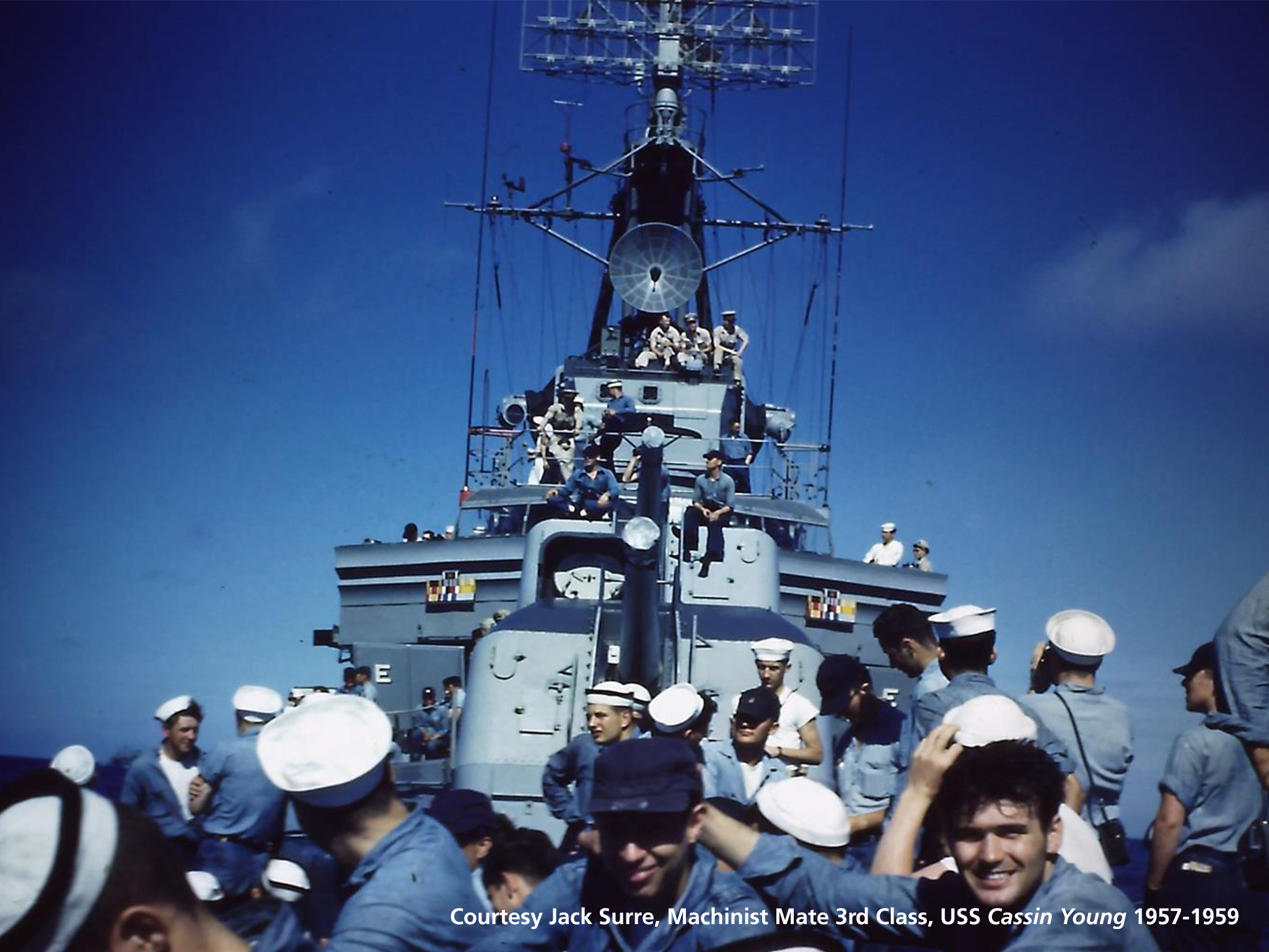 Color photograph of men in blue uniforms and white hats standing on the bow of the ship. 
