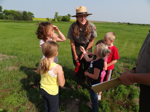 Female ranger talks with 5 children outside on the grass. 