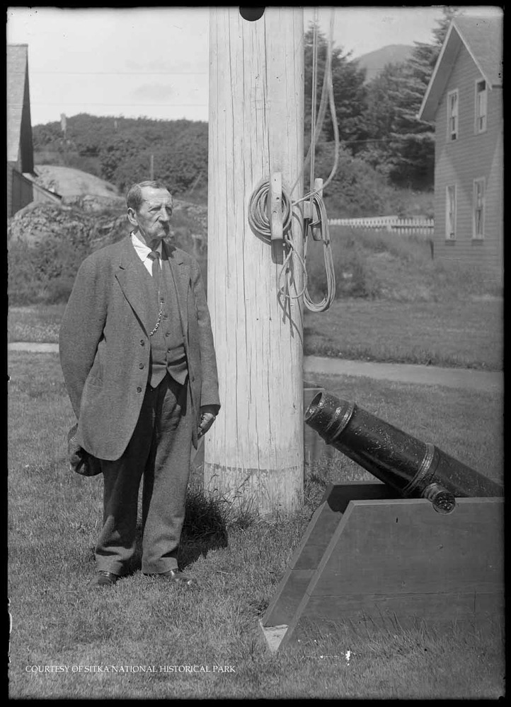 Peter Chernoff posing by a flag pole and small cannon. 1913.