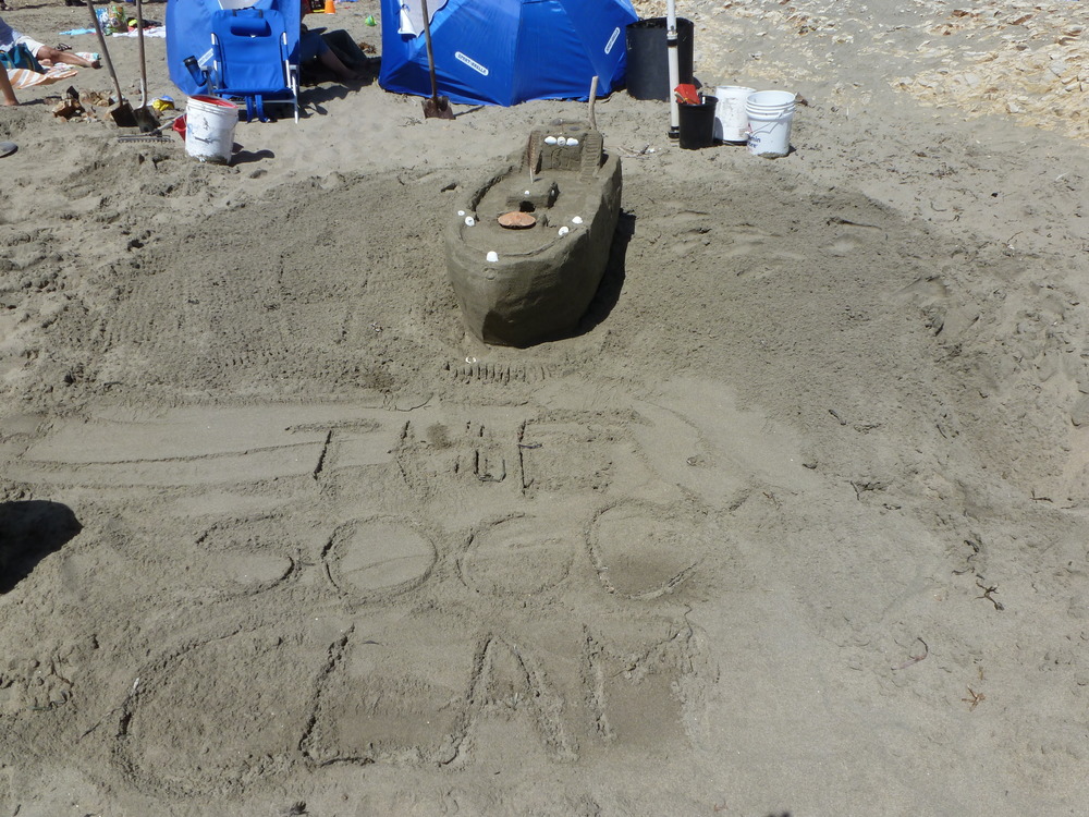 A sand sculpture of a boat with the words 'The Soggy Clam' written in the sand in the foreground.