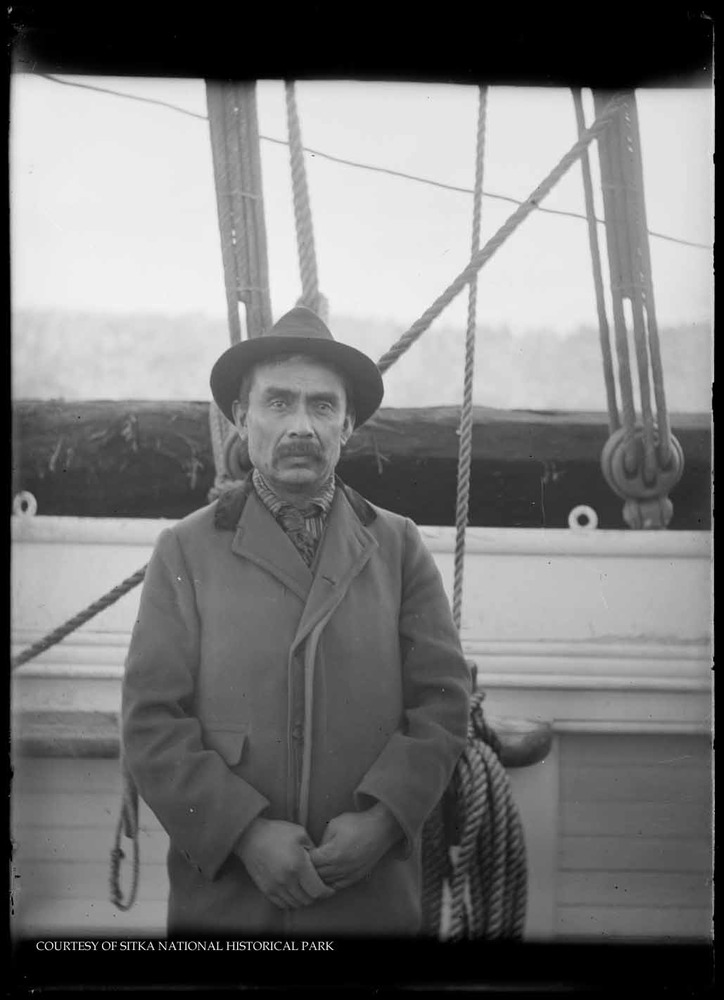 Alaska Native man wearing a coat and hat and standing on board a boat.