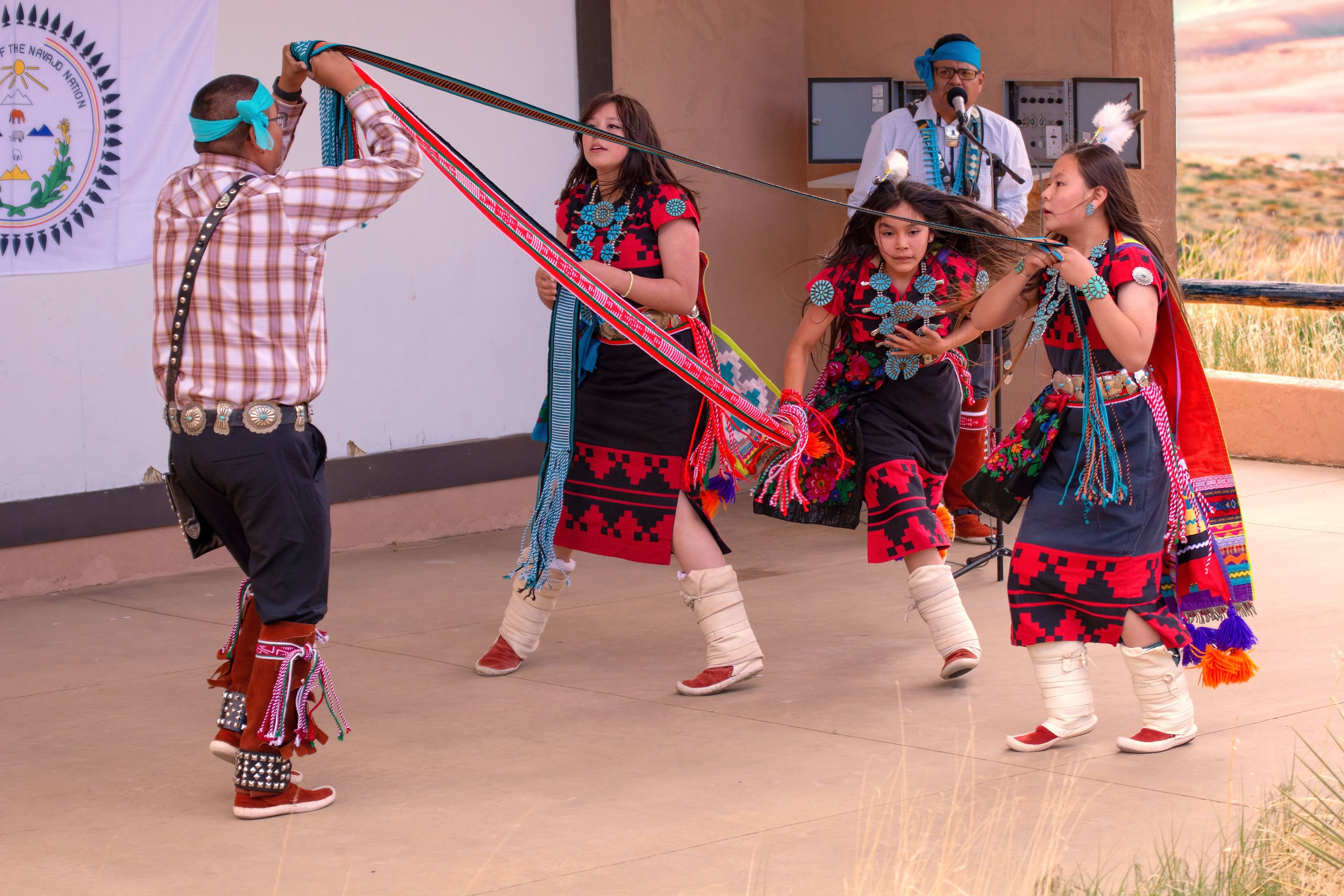 Three Dineh Tah' Navajo Dancers weave a long sash as they dance during a presentation at the park Amphitheater. They are wearing colorful traditional black and red dresses and turquoise jewelry. A Navajo man holds one end of the sash while another sings and plays hand drum.