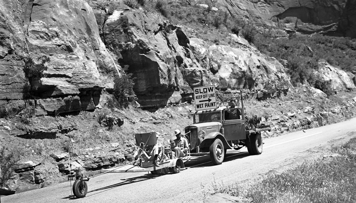 Three men operate truck and machine for painting center line stripe on road in Zion Canyon. Sign of truck says: Slow / Keep Off Line / Wet Paint.