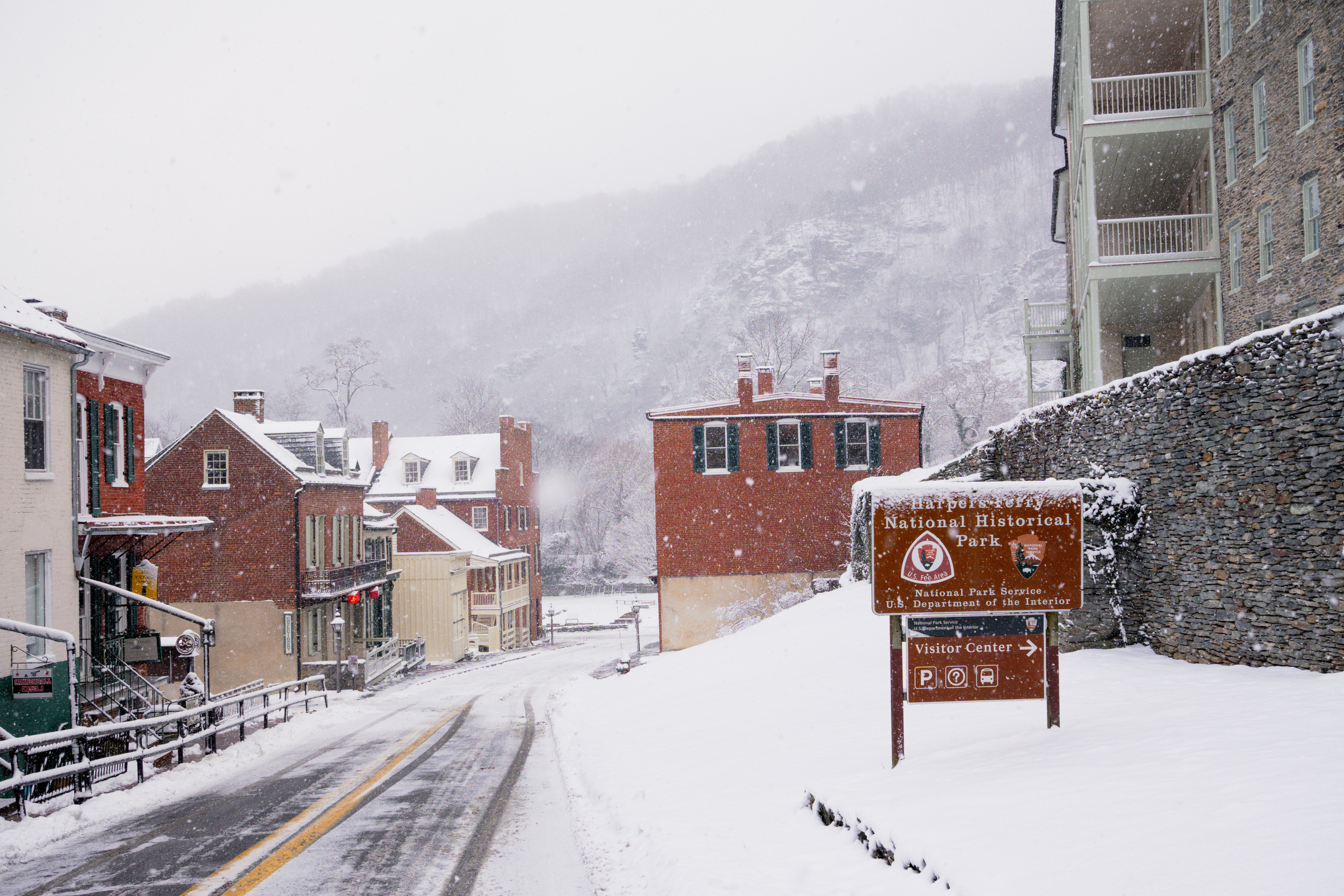 Heavy snowfall begins to cover an entrance sign to Harpers Ferry National Historical Park in West Virginia, with the buildings of Lower Town behind it. In the background, the Blue Ridge is barely discernable.