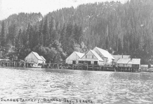 A framework of wooden docks and piers surrounds the cannery structures, an assortment of wooden structures with peaked roofs. A tree-covered hillside rises in the background. Black and white photo; hand-printed text at bottom says: "Dundas Cannery, Dundas Bay, Alaska."