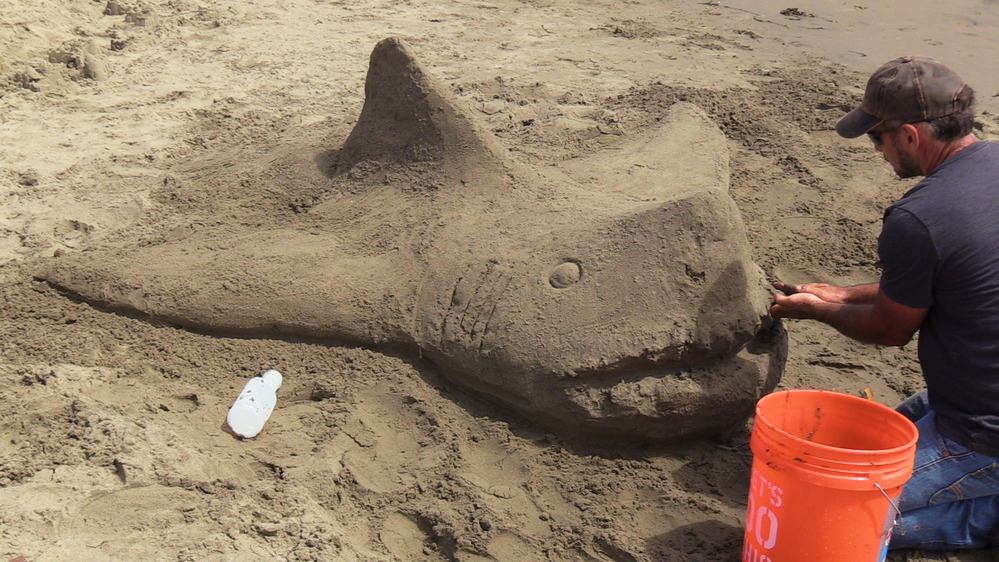 A man working on a sand sculpture of a shark.