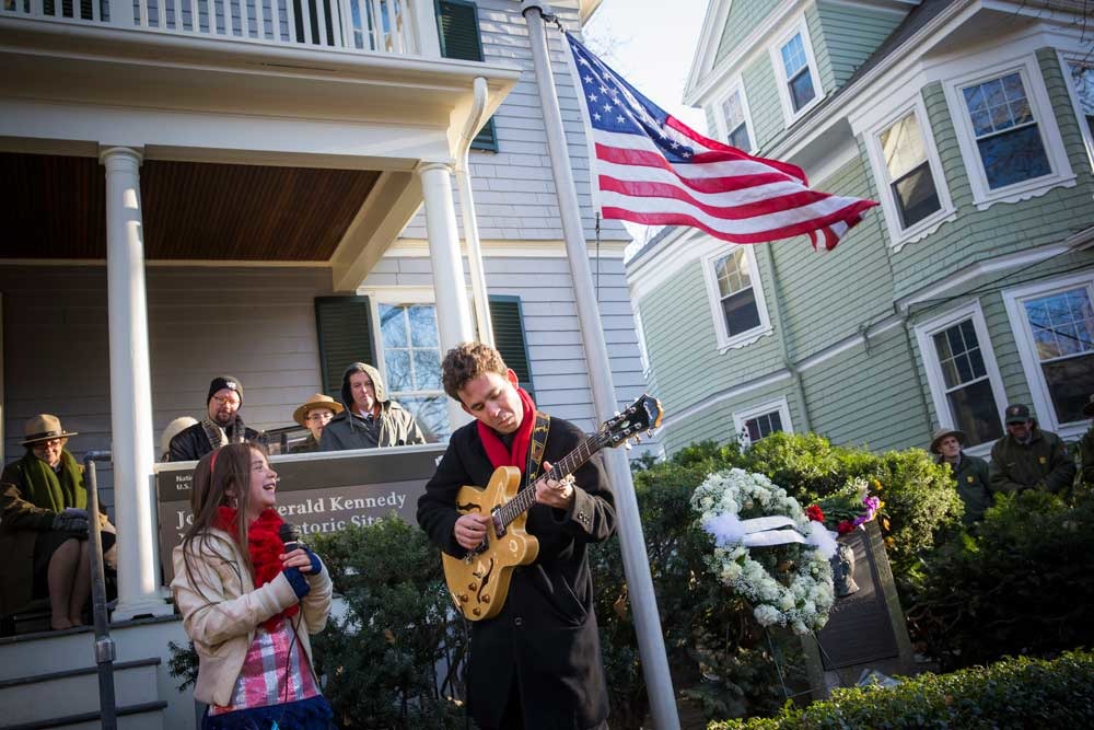 A girl sings while a man play guitar in front of the John F. Kennedy National Historic Site