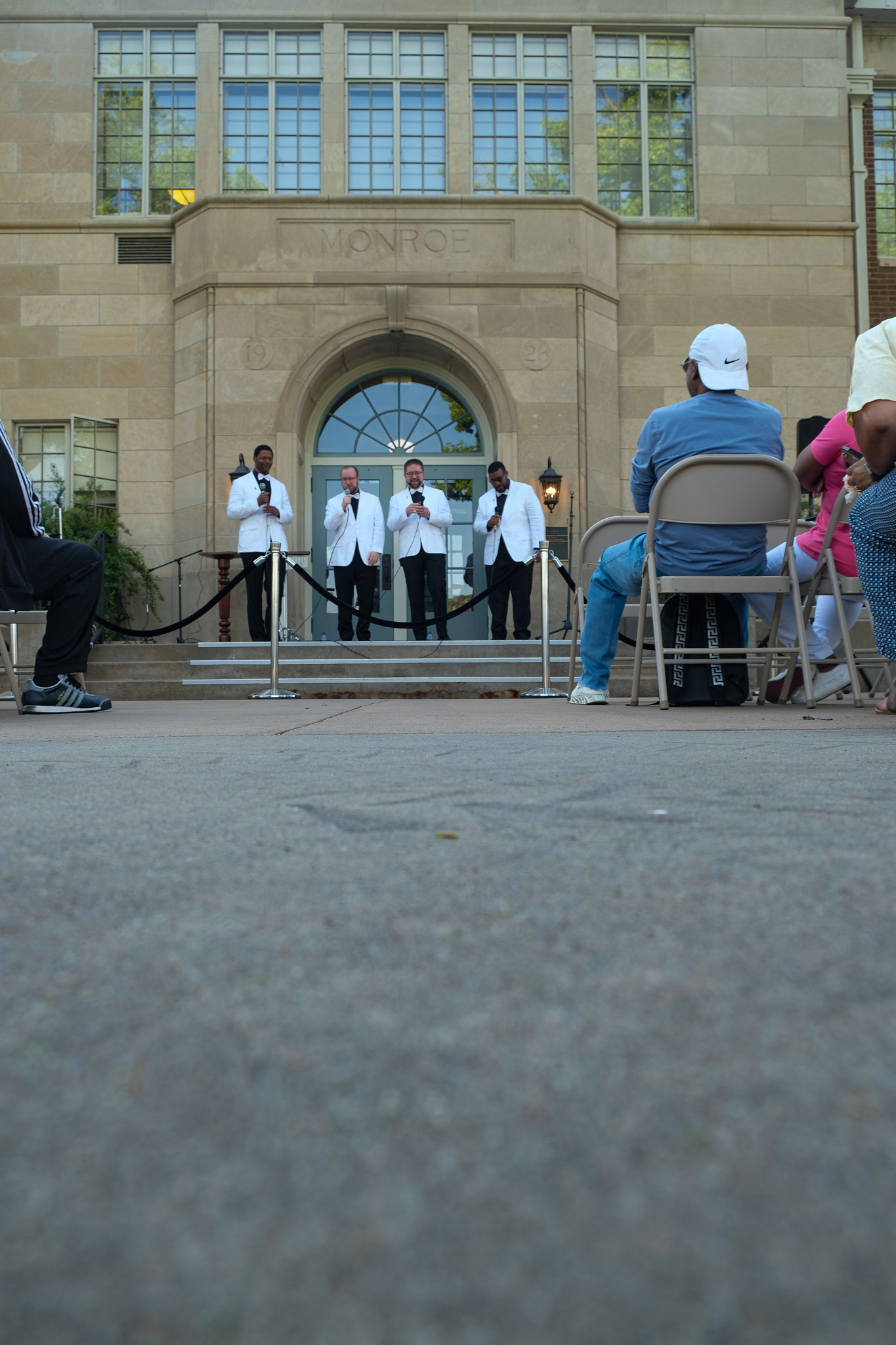 Two African American men and two White men can be seen singing together as a quartet in the center of the image. A crowd of seated onlookers can be seen on the left and right side of the image. An early 20th century school is visible in the background. An asphalt path is visible in the foreground.