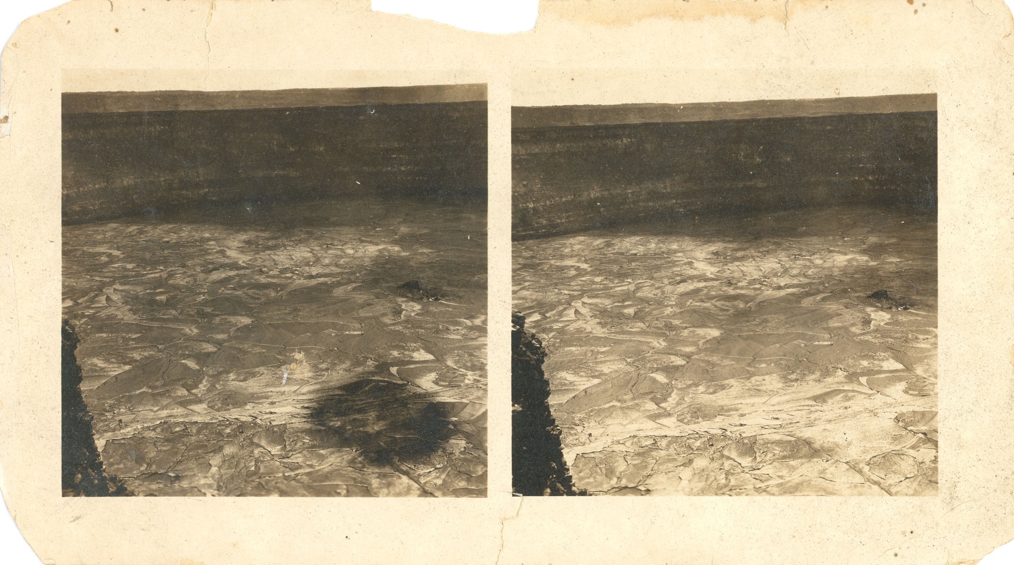 Two nearly identical black and white photographs mounted on brown paperboard. The images depict the vast interior of a crater with smooth lava. The photograph on the left side appears to be taken moments before or after the photograph on the right, as evidenced by the shadow of a small cloud and a slightly different perspective. The wall of the crater is seen in the background.
