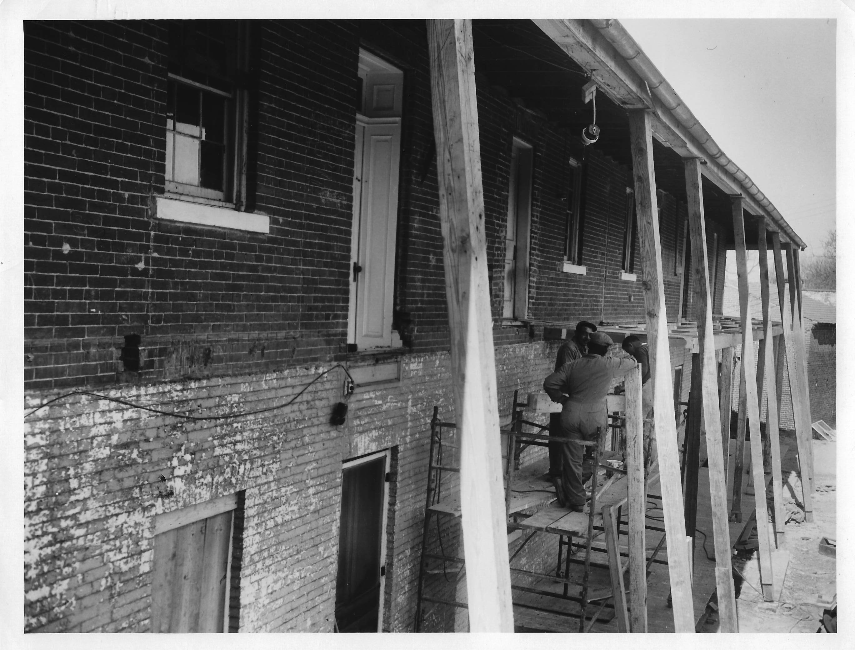 The back of the Officer's Quarters.  The roof of the porch is being supported by beams secured at the ground.  The porch is partially rebuilt there are two workman on scaffolding.  
The coloring on the brick is two-toned, darker on the upper floor and lighter on the lower floor.  