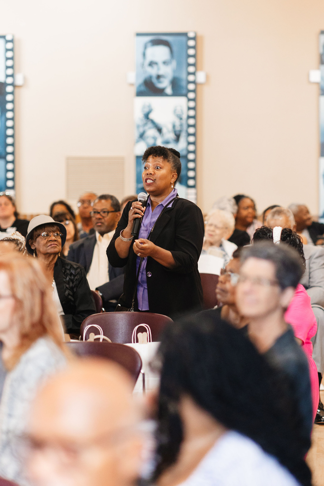 an african american woman in a black jacket and purple shirt stands while speaking into a microphone while surrounded by an audience of mostly african american adults seated in maroon plastic chairs
