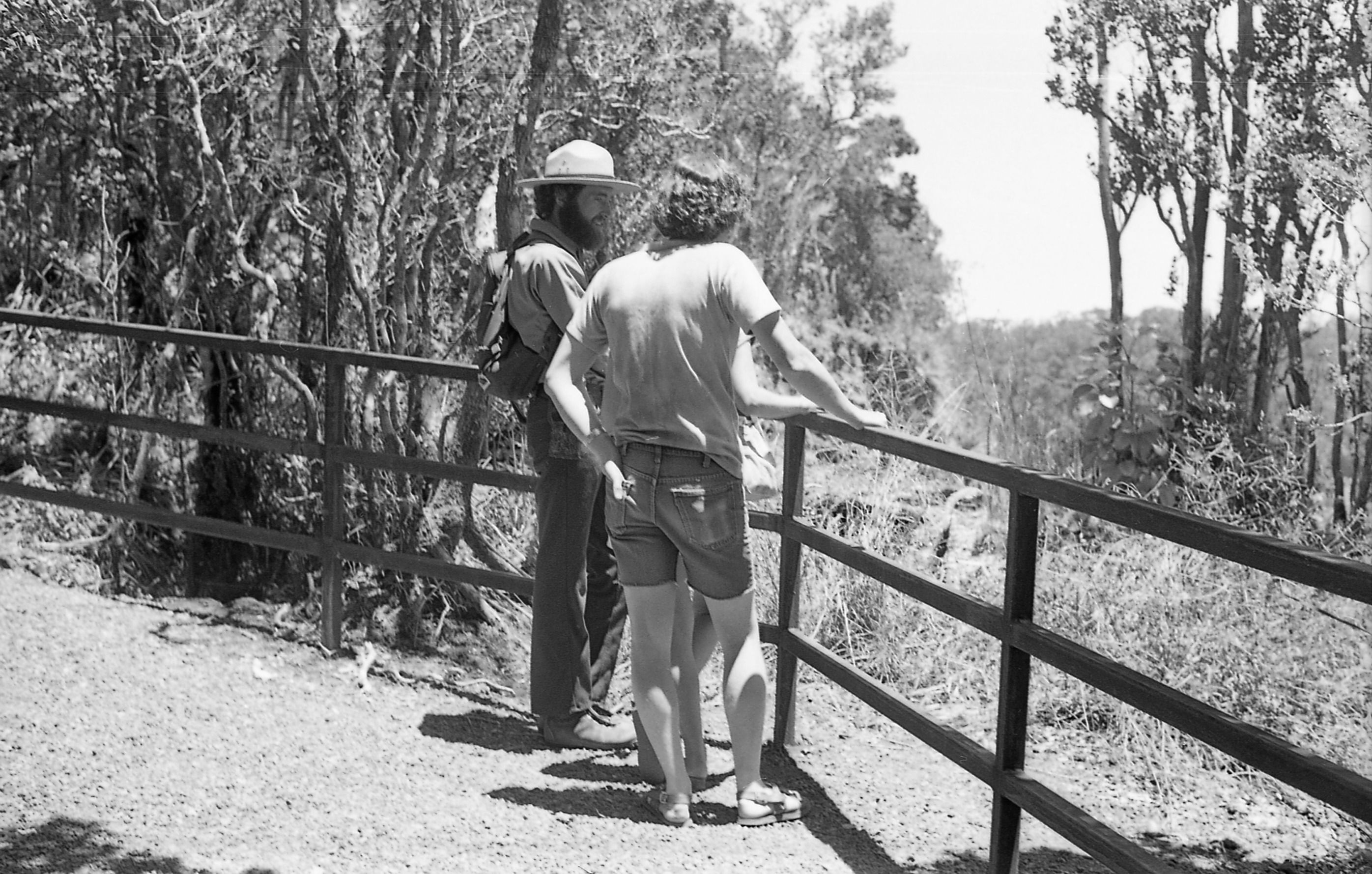 A black and white image of a male park ranger talking to two visitors on the side of a trail. The park ranger and visitors are standing next to a wooden railing in the center of the image. The park ranger is wearing a ranger hat, a long sleeve collared shirt, pants, and boots. He is carrying a backpack slinged across his back. To the right of him is a visitor who we can only see their legs and right arm. Lastly, behind this visitor is a man with curly hair. He is wearing a t-shirt, cut-off jean shorts, and sandals. His right hand is holding onto the top of the wooden railing. In the background behind the railing is a wooded landscape.