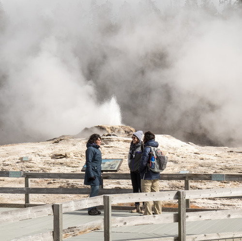 Three people in warm clothes standing in front of an erupting geyser talking with one another