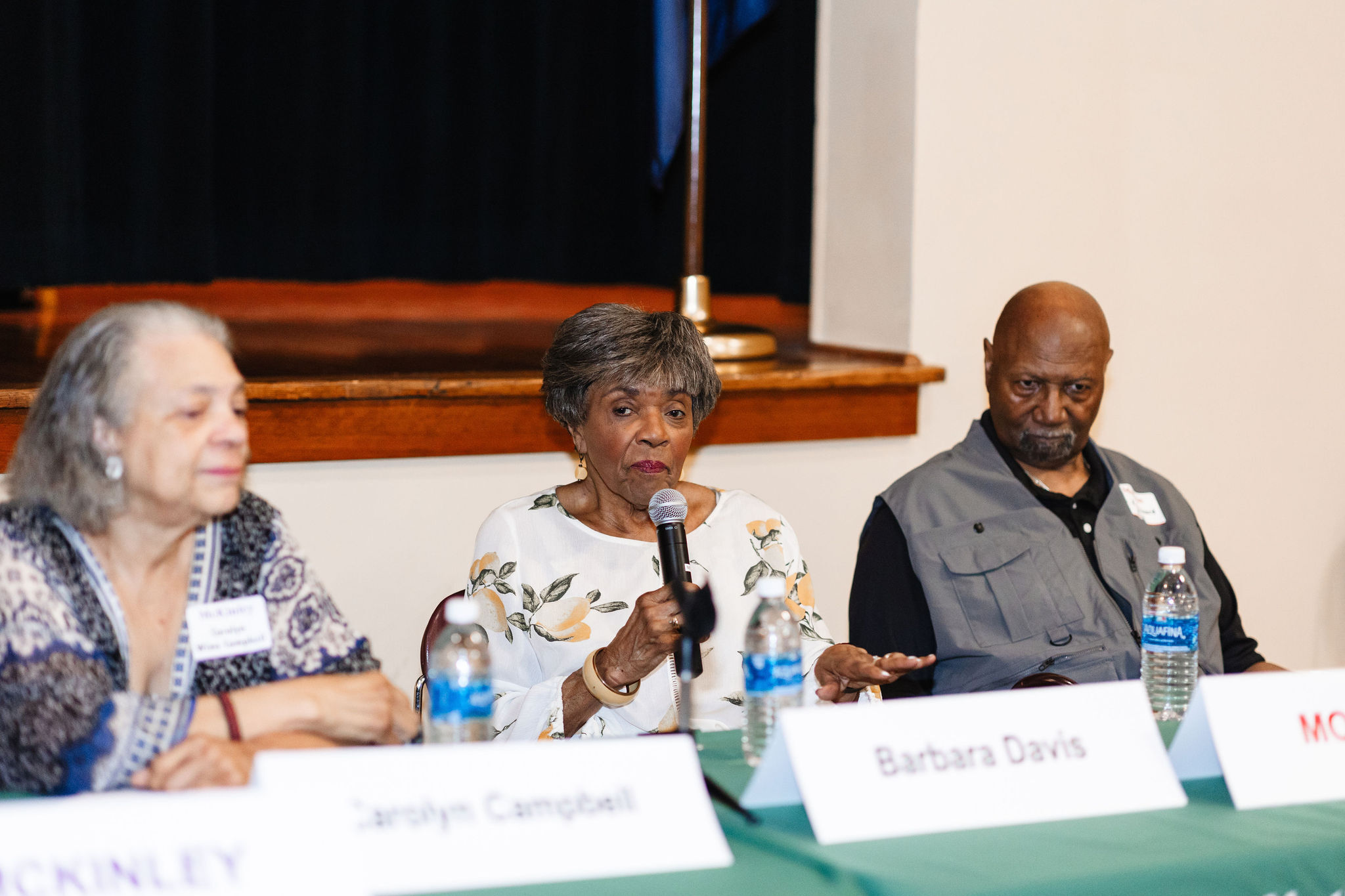 An older african american panelist in a white shirt with lemons and leaves on it speaks into a microphone while seated behind a green table and a table tent reading barbara davis