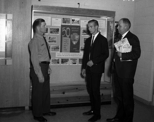 Superintendent Warren F. Hamilton and Jim Clarke stand with Creed Fraehner in front of Fraehner's winning poster exhibit for the National Park Service 50th Anniversary, inside the Mission 66 Visitor Center and Museum.