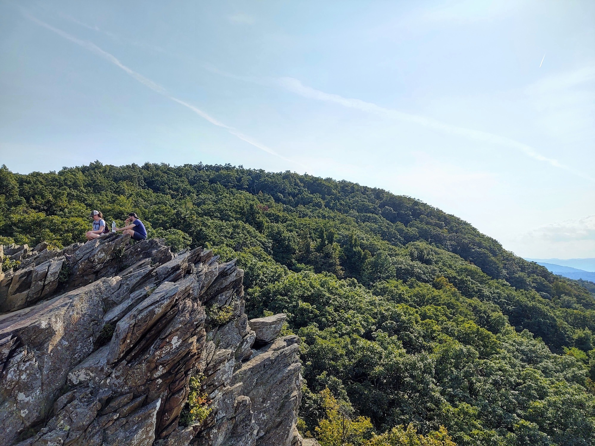 Two hikers sitting on top of a large rock with a tree covered mountain as the view. 