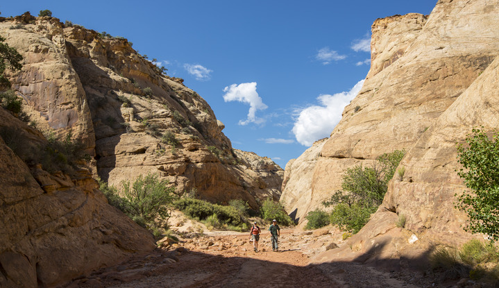 Two hikers in a deep canyon, with tan cliffs on either side, and blue sky with clouds above. 