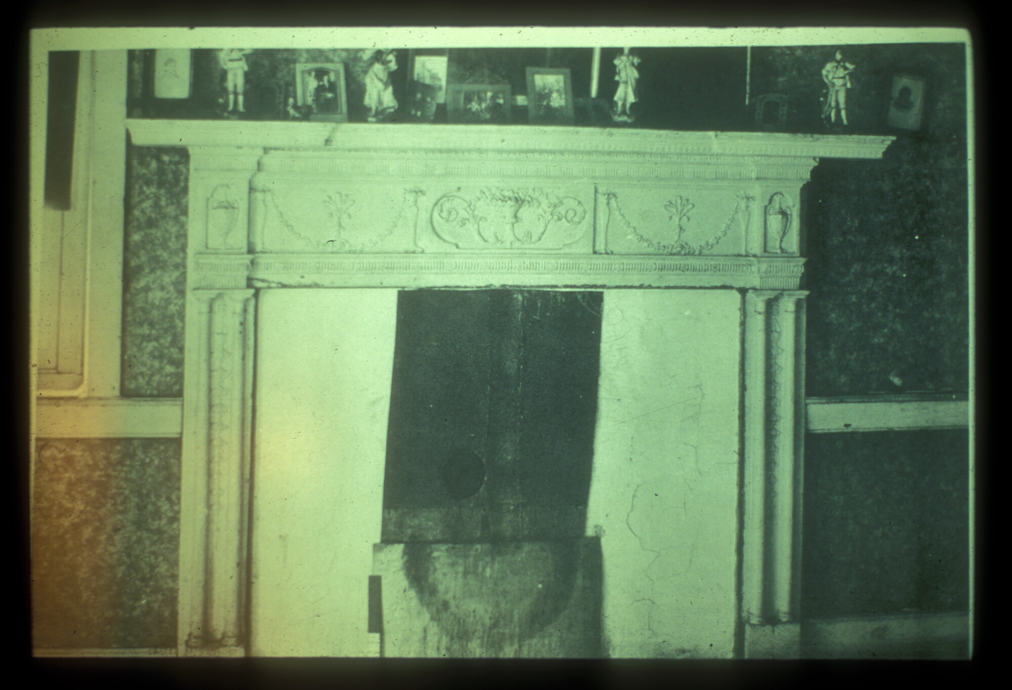 A highly decorated/molded mantle piece with small items on the shelf. Textured wallpaper is visible in the background.