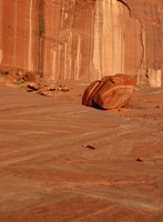 Photograph of steep orange-and brown sandstone cliff with prominent streaks of dark desert varnish. Cross beds are well defined. Large boulders have fallen from the cliff onto the slickrock surface.