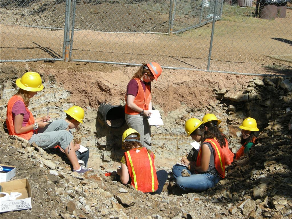 a group of young people sit in a trench wearing safety vests and hard hats while taking notes. 
