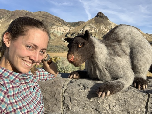A smiling artist-in-Residence, Catherine Danae, poses with a felted wool oreodont with Sheep Rock in the background.