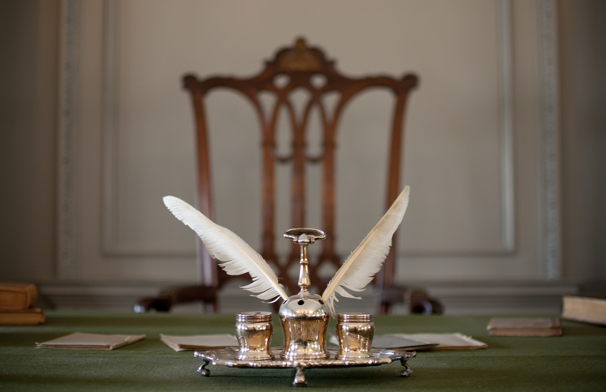 A three piece silver inkstand holding two quill pens sits on a silver tray in the foreground.  The Rising Sun Chair is visible behind the inkstand.