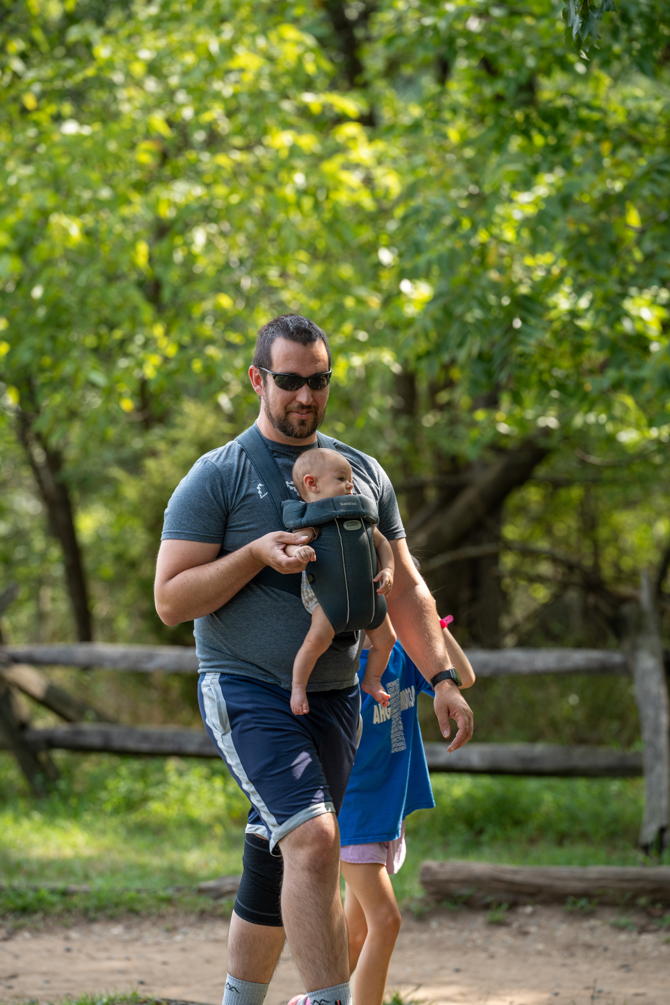 A man with a baby in a baby carrier on his chest walks with another child beside him on a dirt trail. 
