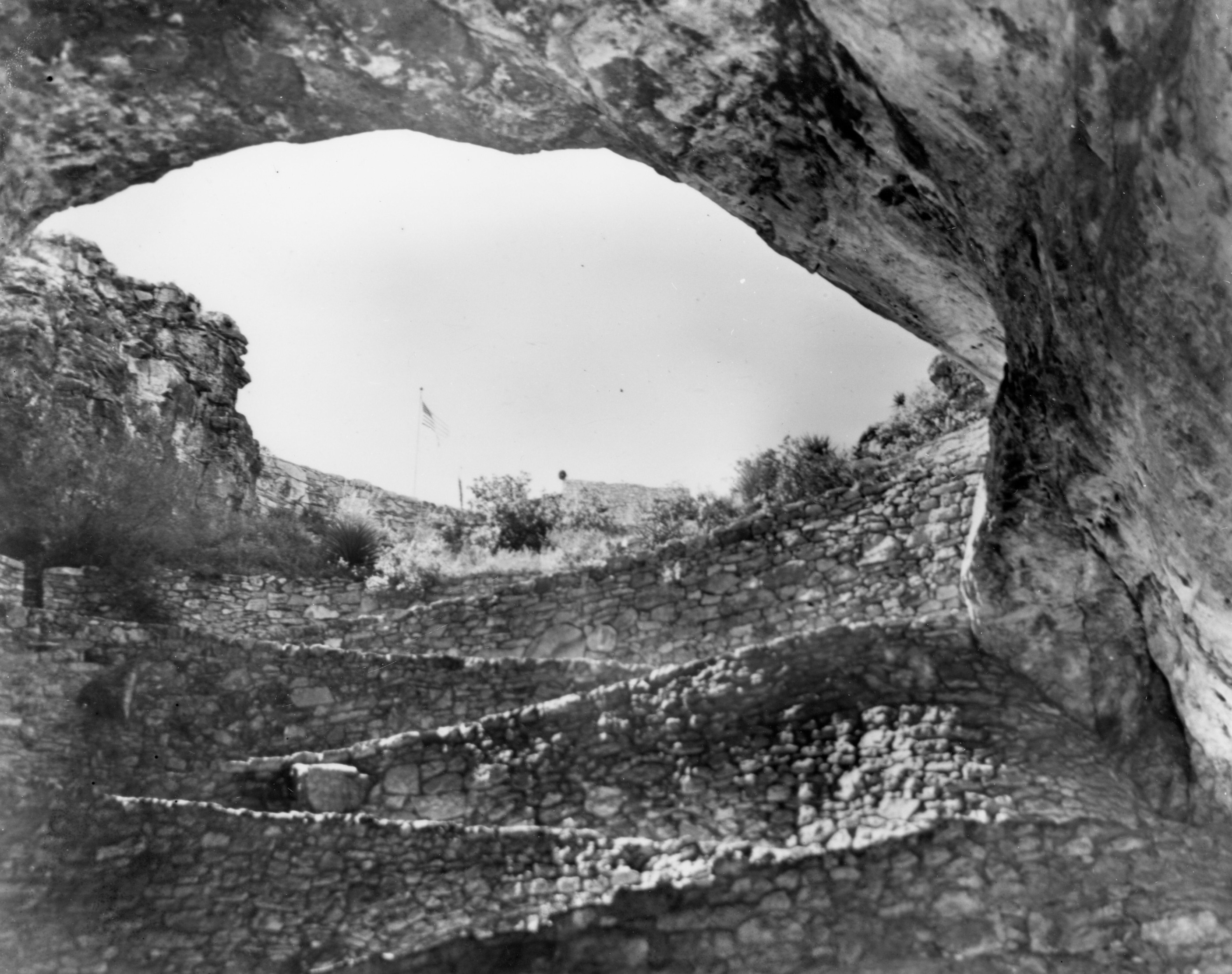 A black and white photograph of the trail leading down from the top of the mouth of the natural entrance. The photograph is looking up at the entrance with the flagpole and United States flag just visible.