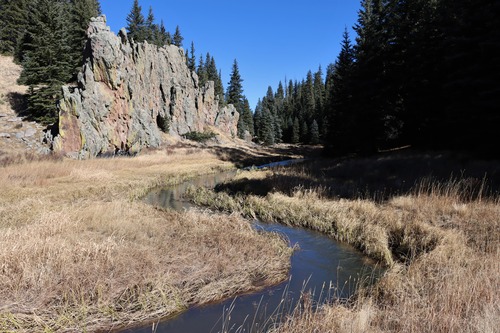 A stream meanders through a narrow valley with spruce trees and rock spires.