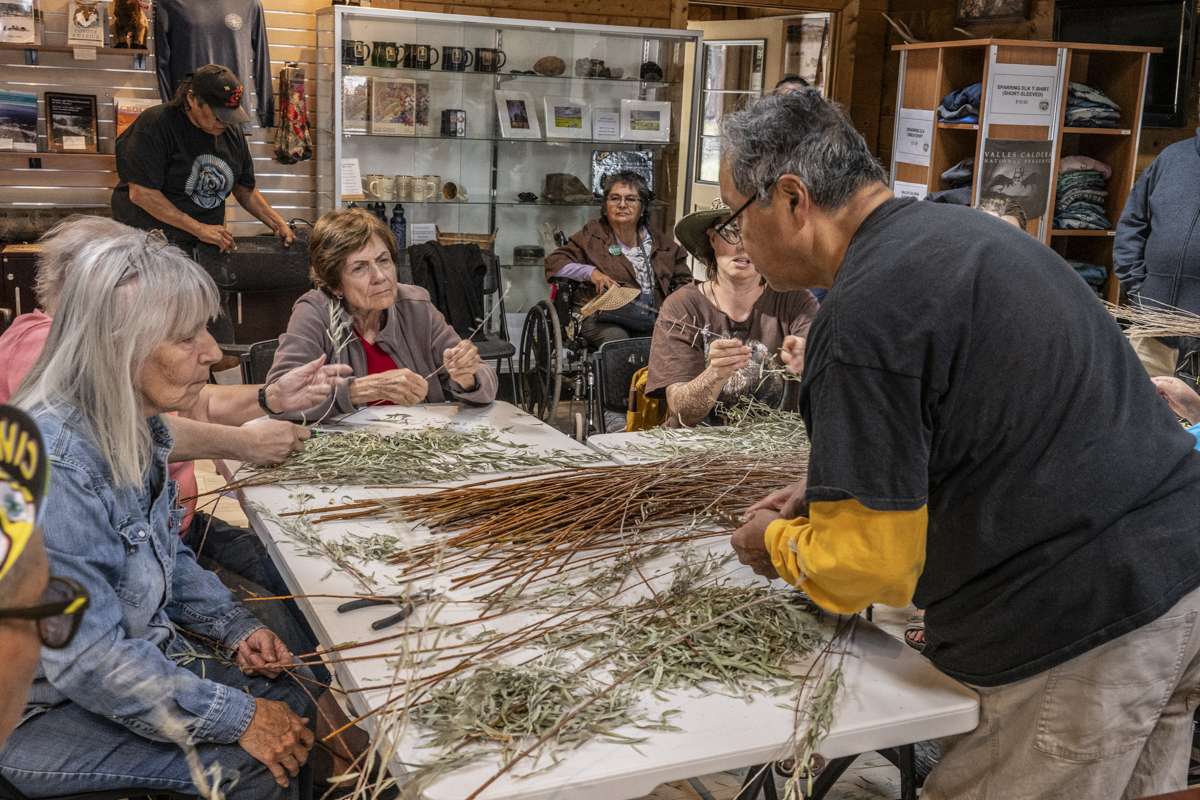 A man stands in front of a seated audience and demonstrates willow basket weaving.