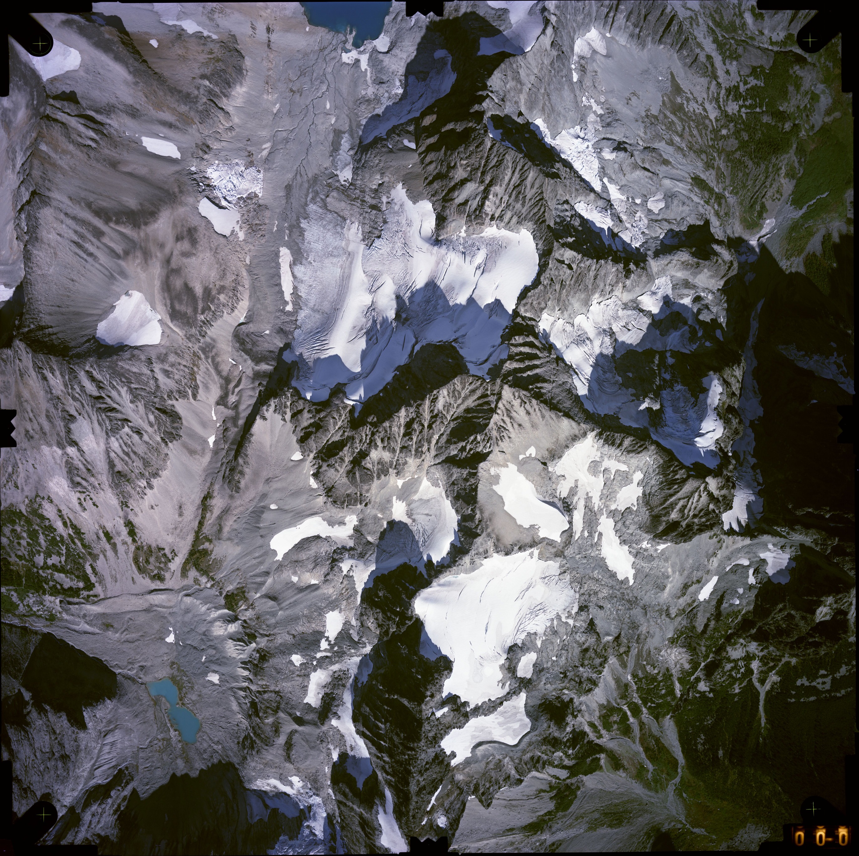 Color aerial photograph of glacier, snowfields, and alpine lake on mountain peak with forested valleys below.